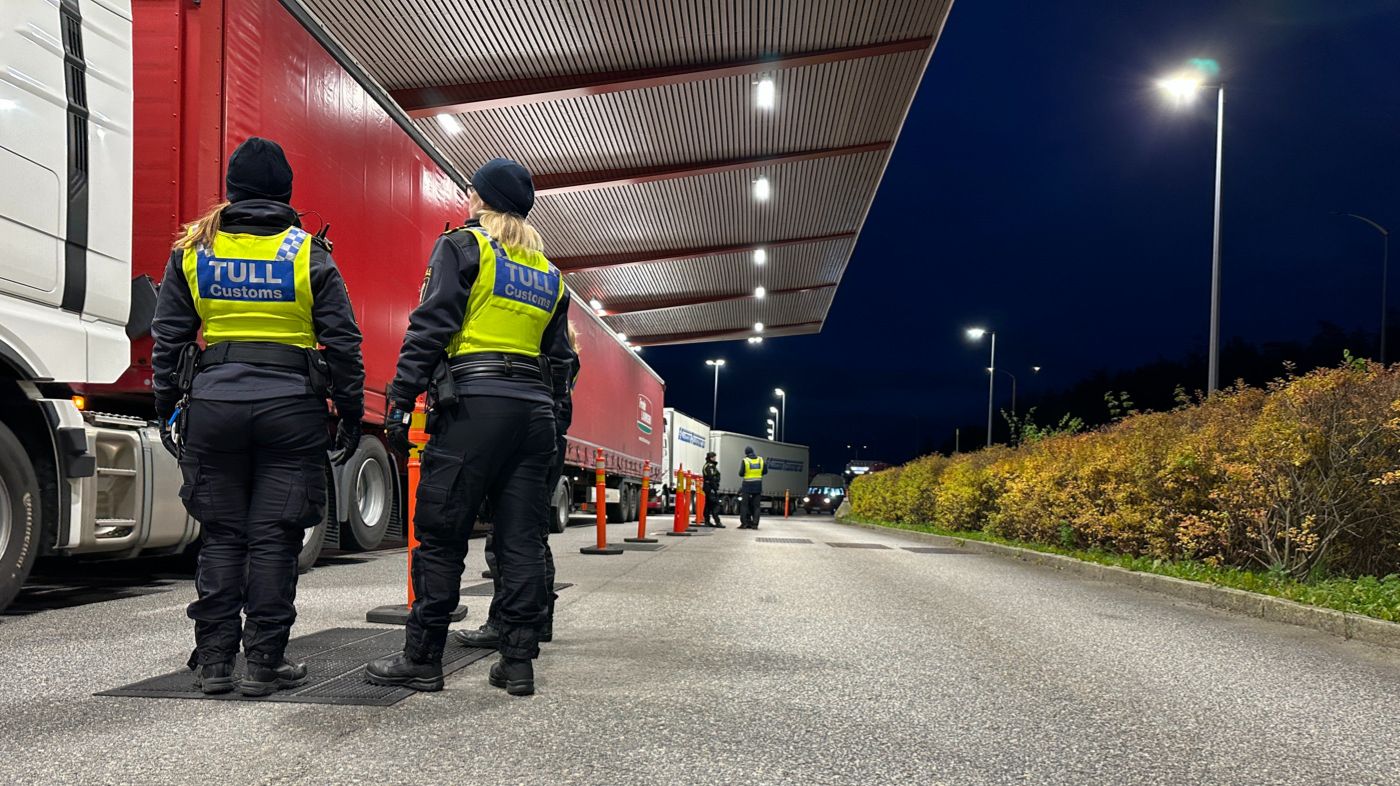Två tullinspektörer står utomhus intill flera lastbilar.